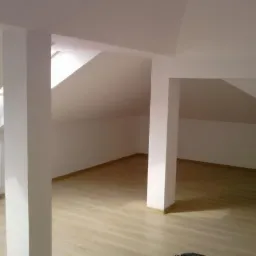 Puste, białe poddasze z drewnopodobną podłogą, widoczne dwie kolumny konstrukcyjne i okno dachowe.