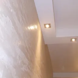 Ściana wewnętrzna z dekoracyjną, perłową fakturą w odcieniach beżu i bieli, oświetlona przez kwadratowe lampy sufitowe wbudowane w podwieszany sufit.