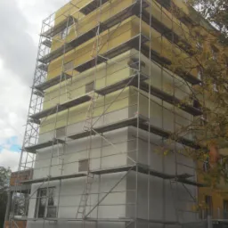 Wielopiętrowy budynek w trakcie docieplania, pokryty rusztowaniem, widoczne fragmenty elewacji w kolorze żółtym i białym, dwóch pracowników na rusztowaniu, a dwóch na dole, jesienne drzewo w tle.