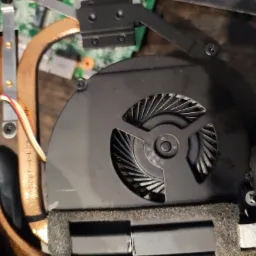 Czyszczenie wentylatora chłodzenia laptopa z widocznymi miedzianymi rurkami cieplnymi i elementami płyty głównej w tle, wykonywane w rękawicach ochronnych.