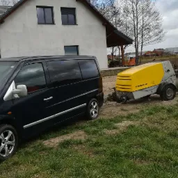 Czarny van Mercedes 108 CDI z przyczepioną maszyną Putzmeister M 740 na trawiastym terenie przed domem z szarą elewacją.