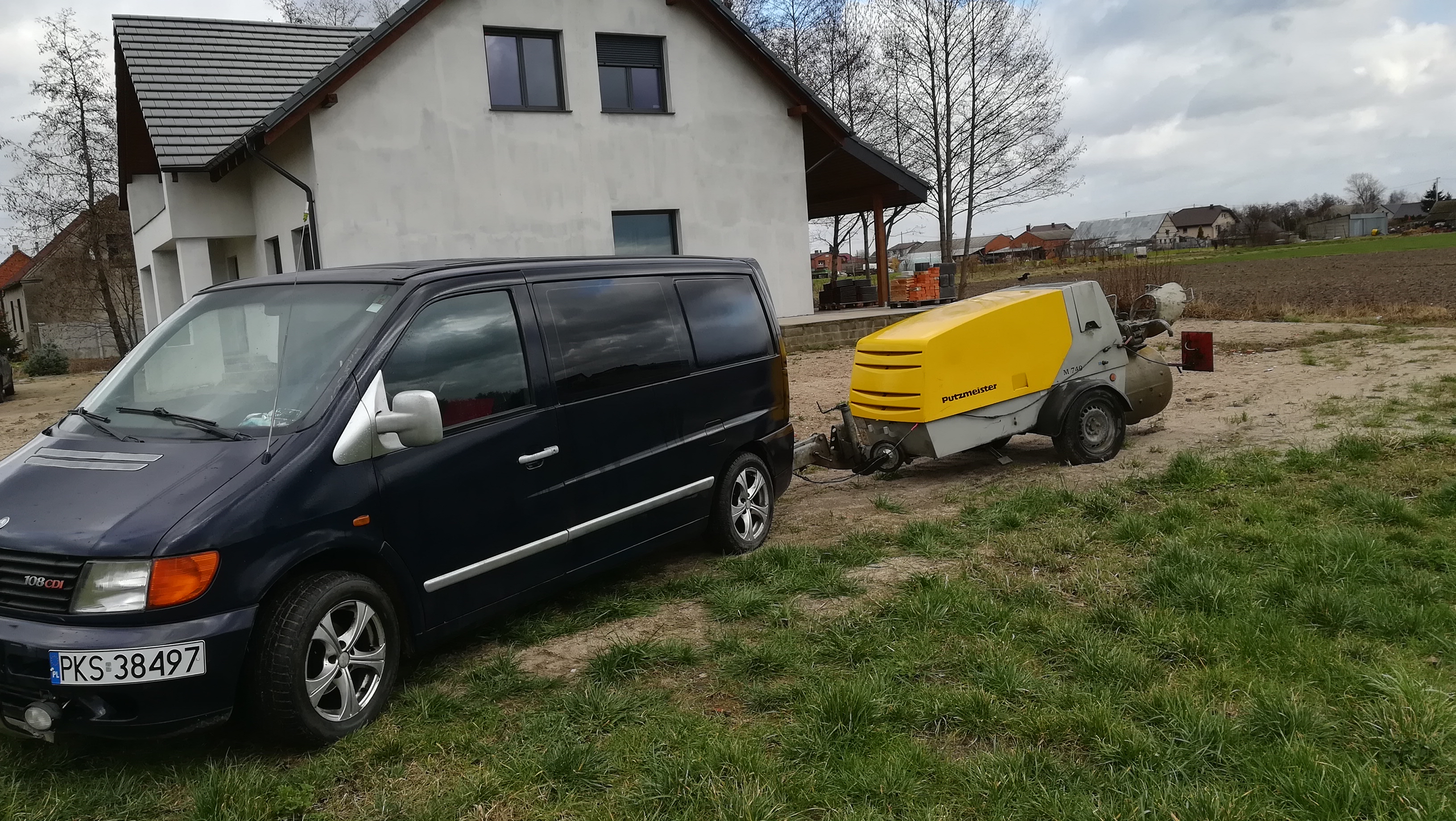 Czarny van Mercedes 108 CDI z przyczepioną maszyną Putzmeister M 740 na trawiastym terenie przed domem z szarą elewacją.