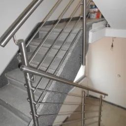 Schody z szarymi, granitowymi stopniami zabezpieczone metalową, minimalistyczną barierką z poziomych prętów. W tle widoczne wiaderka z farbą.