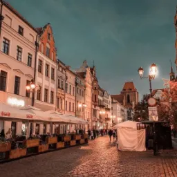 Ulica w starym mieście o zmierzchu, z brukowaną drogą, oświetlona latarniami i parasolami restauracyjnymi, z widoczną wieżą kościoła w oddali.