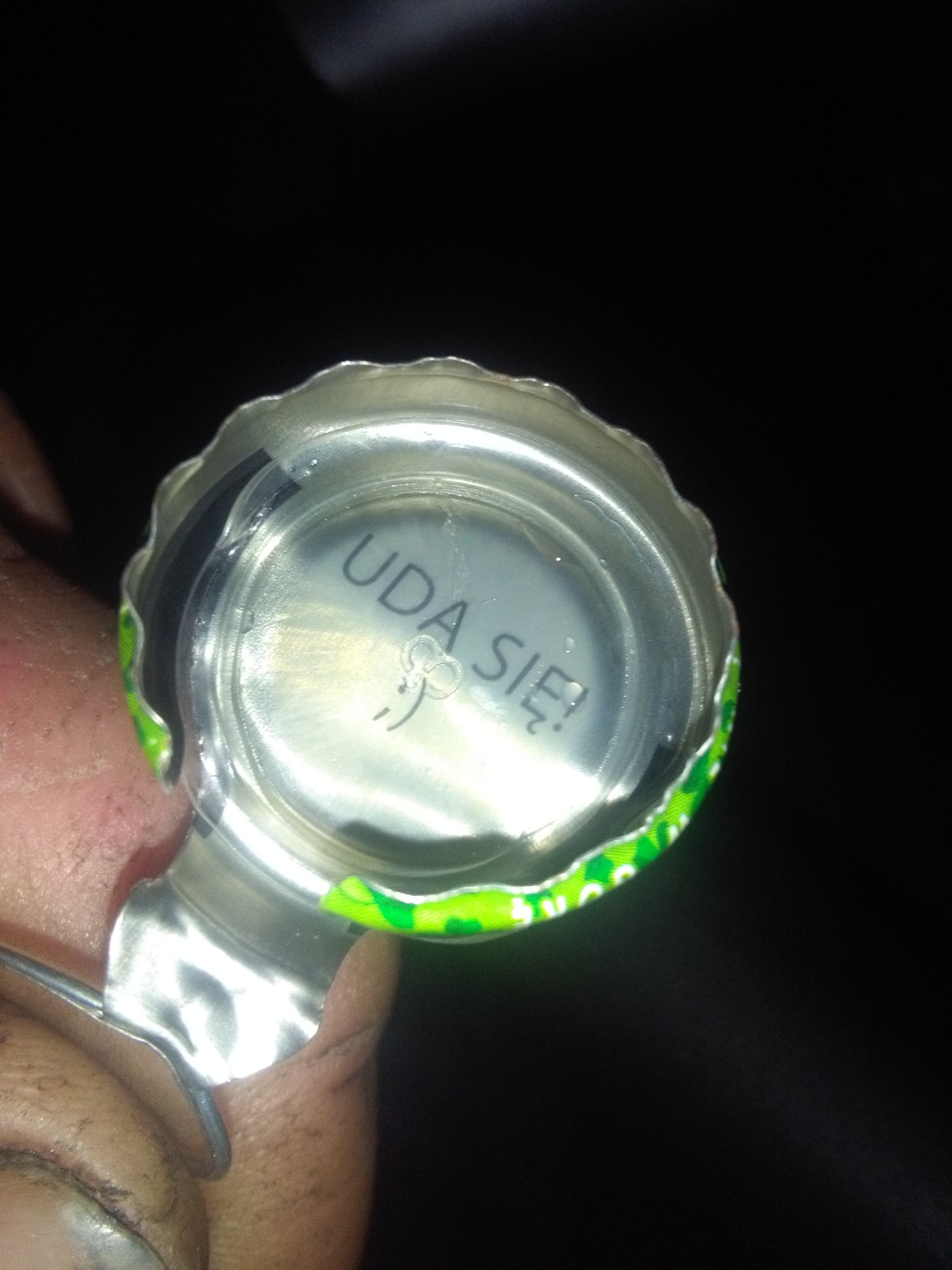 Wnętrze kapsla od napoju z napisem 'UDA SIĘ!' i małą buźką, trzymane w palcach