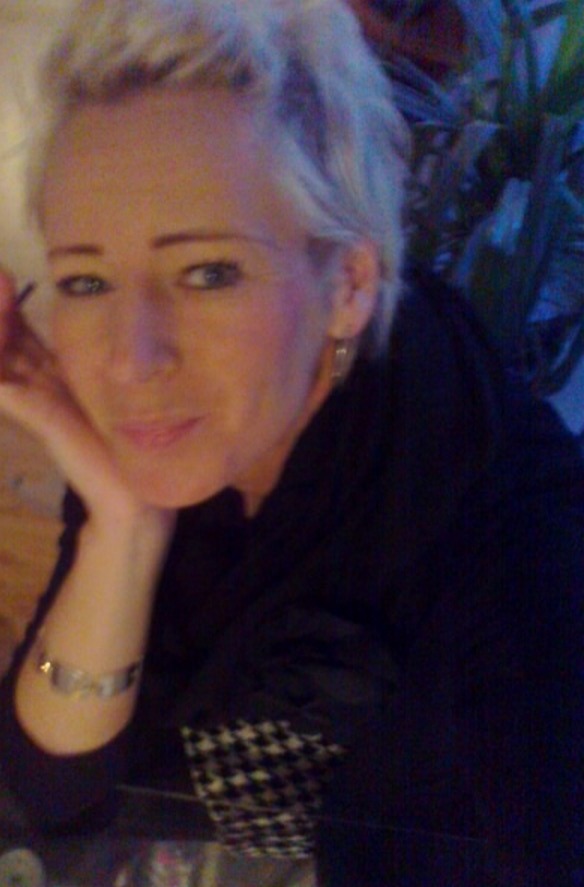 Portret kobiety z krótkimi, siwymi włosami, opierającej brodę na dłoni, z rośliną w tle.