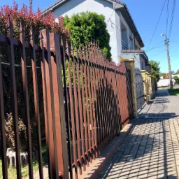 Nowoczesne, brązowe ogrodzenie metalowe z dekoracyjnymi grotami, przylegające do chodnika z cieniem rzucanym przez płot, na tle jasnoszarego domu z brązowym dachem i zielenią.