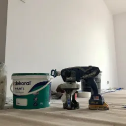 Wnętrze pokoju w trakcie prac przygotowawczych do malowania, widoczny grunt Dekoral, bezprzewodowy agregat malarski Graco Ultra oraz zabezpieczona podłoga.