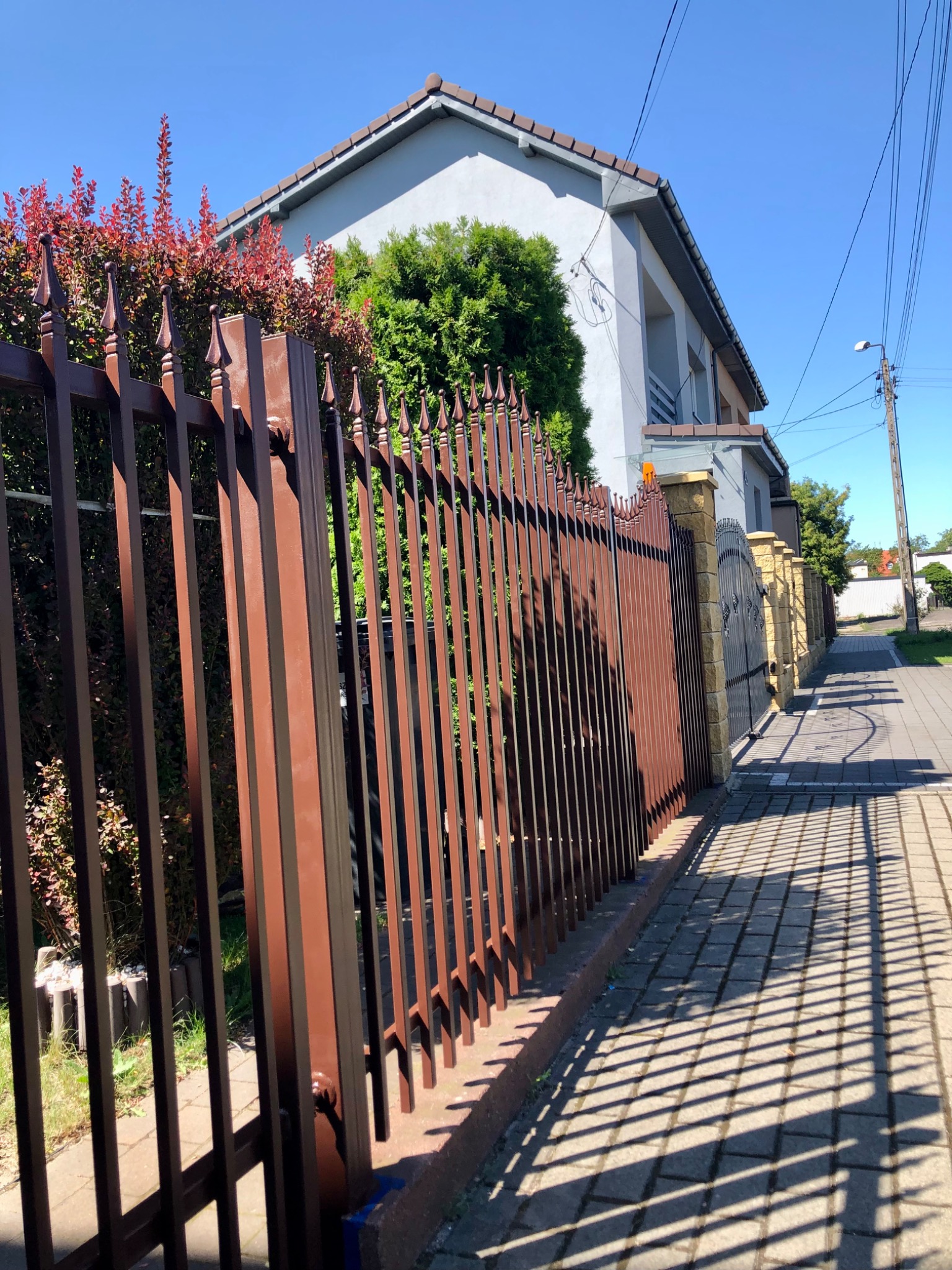 Nowoczesne, brązowe ogrodzenie metalowe z dekoracyjnymi grotami, przylegające do chodnika z cieniem rzucanym przez płot, na tle jasnoszarego domu z brązowym dachem i zielenią.