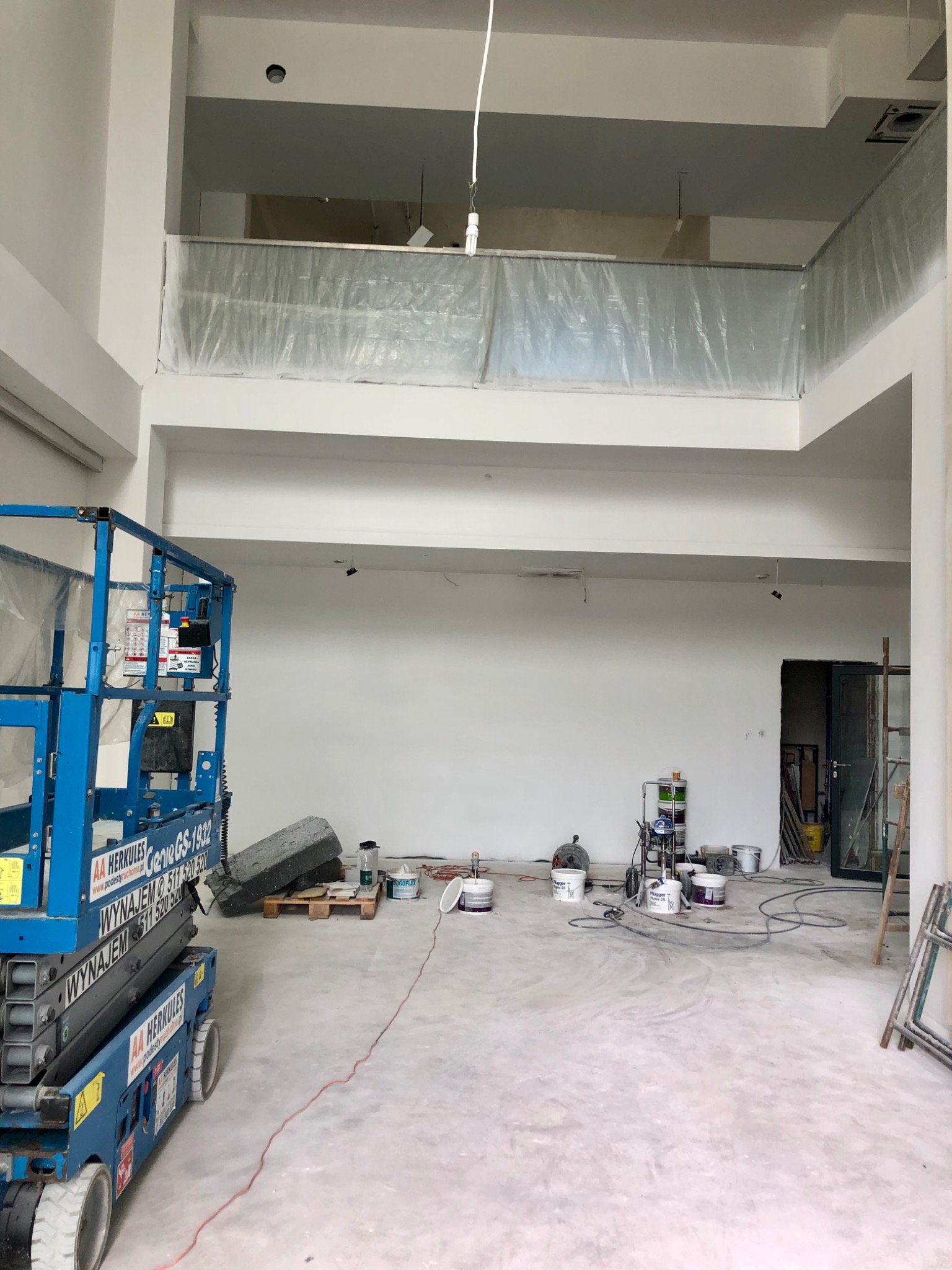 Wnętrze biura w trakcie malowania z podnośnikiem Herkules, wiaderkami farb i zabezpieczonym balkonem na wyższej kondygnacji.