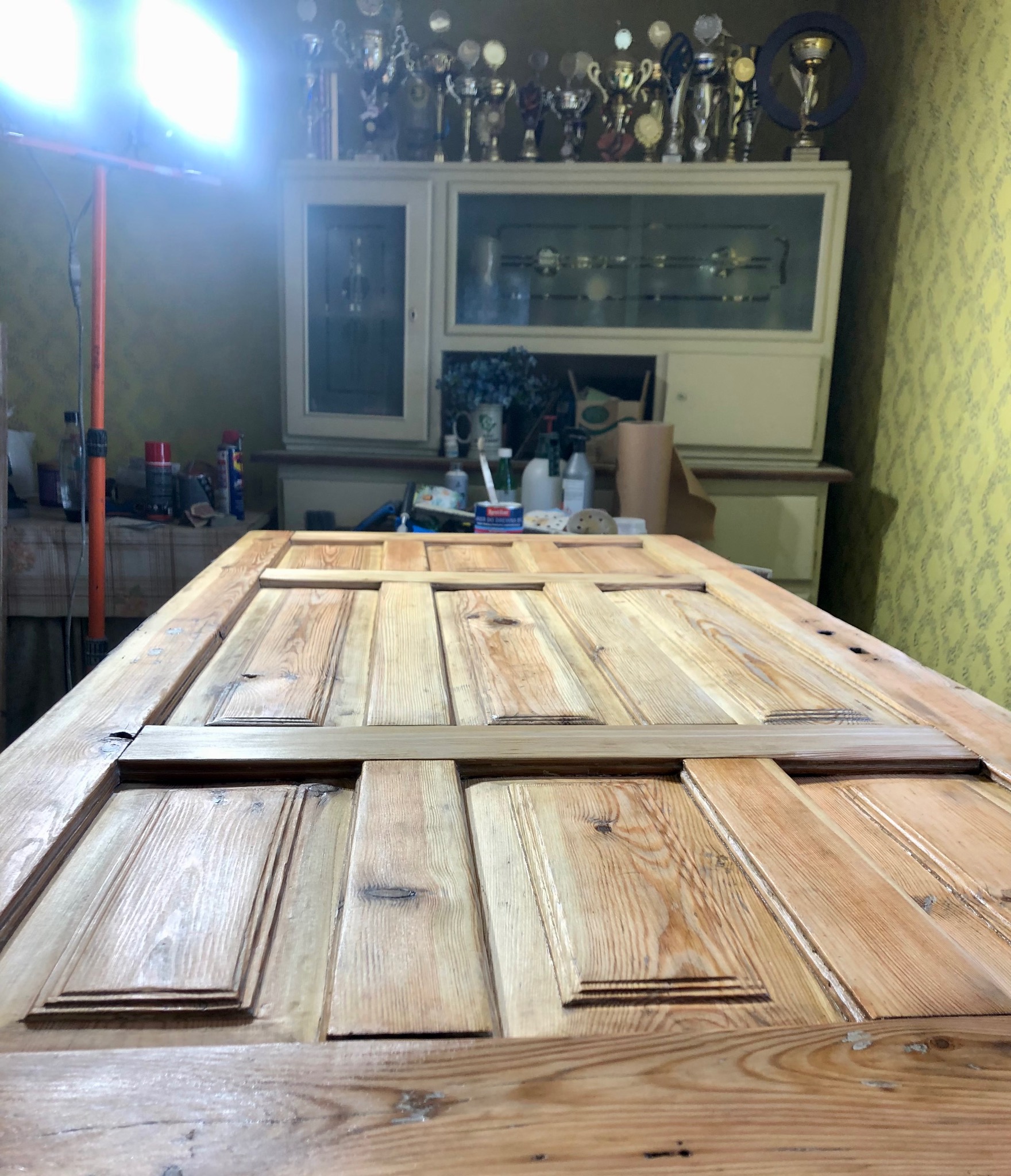 Odrestaurowane drewniane drzwi z widoczną strukturą drewna, leżące na stole w warsztacie. W tle szafka z trofeami i narzędzia.