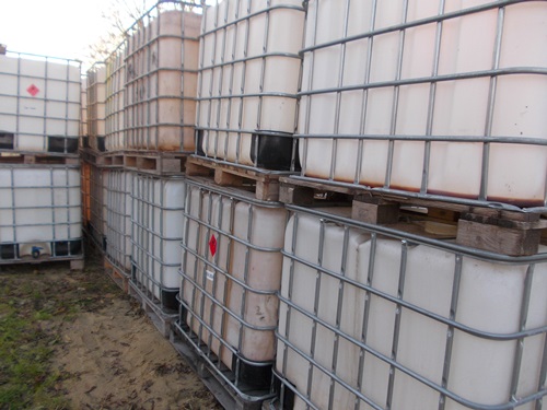 Stos paletopojemników IBC 1000l na drewnianych paletach, niektóre z oznaczeniami substancji niebezpiecznych, widoczne ślady użytkowania.