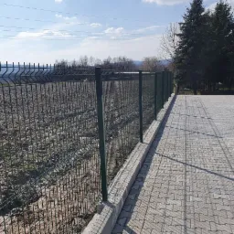 Zielona siatka ogrodzeniowa panelowa na betonowej podmurówce, oddzielająca utwardzony plac z kostki brukowej od przygotowanego terenu pod uprawę, w tle drzewa i błękitne niebo.