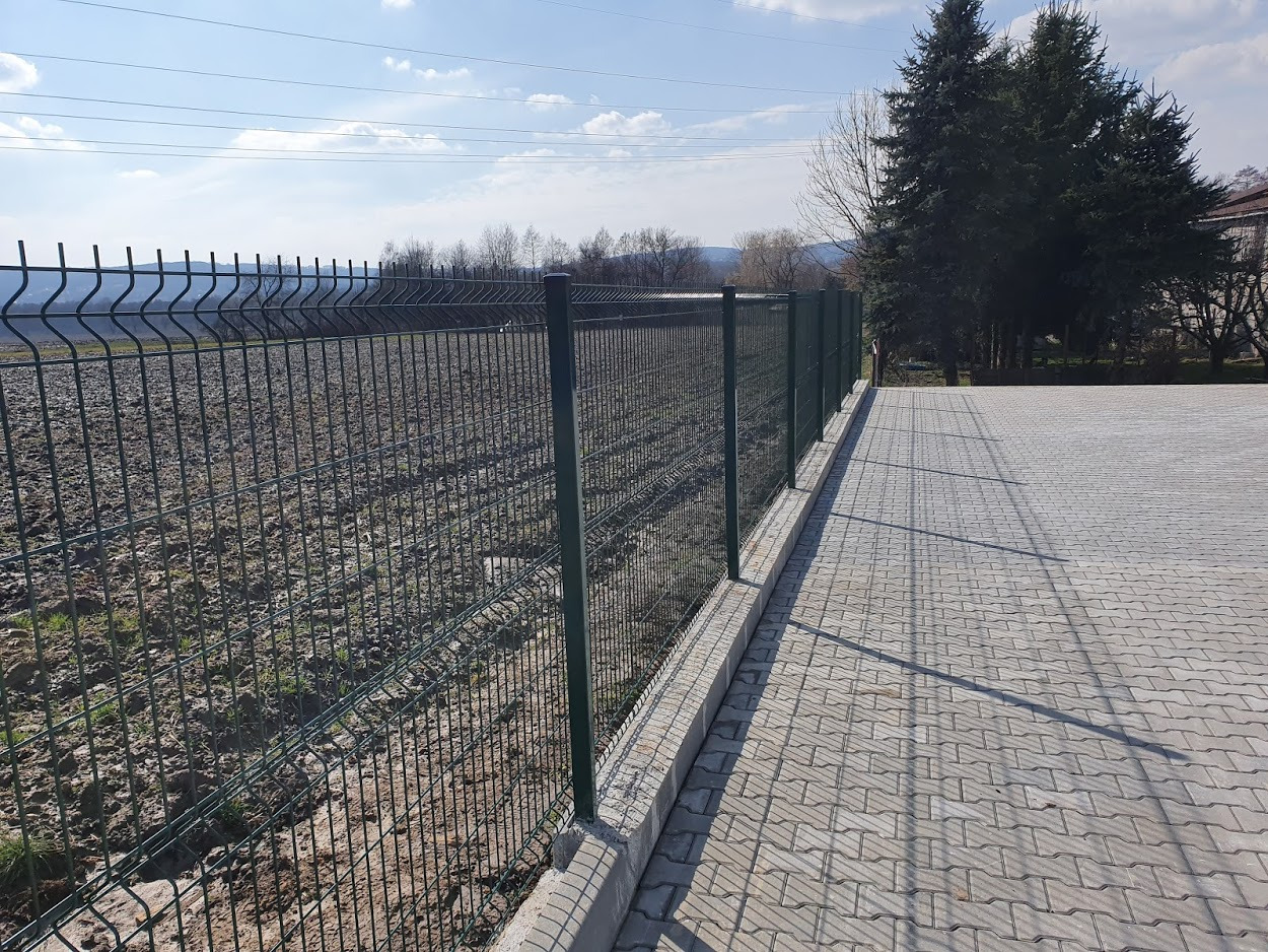 Zielona siatka ogrodzeniowa panelowa na betonowej podmurówce, oddzielająca utwardzony plac z kostki brukowej od przygotowanego terenu pod uprawę, w tle drzewa i błękitne niebo.