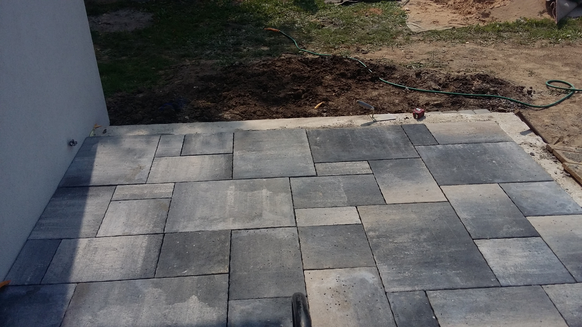 Ułożona kostka brukowa w odcieniach szarości przy ścianie budynku, z widocznym fragmentem ziemi i trawy w tle oraz zielonym wężem ogrodowym.