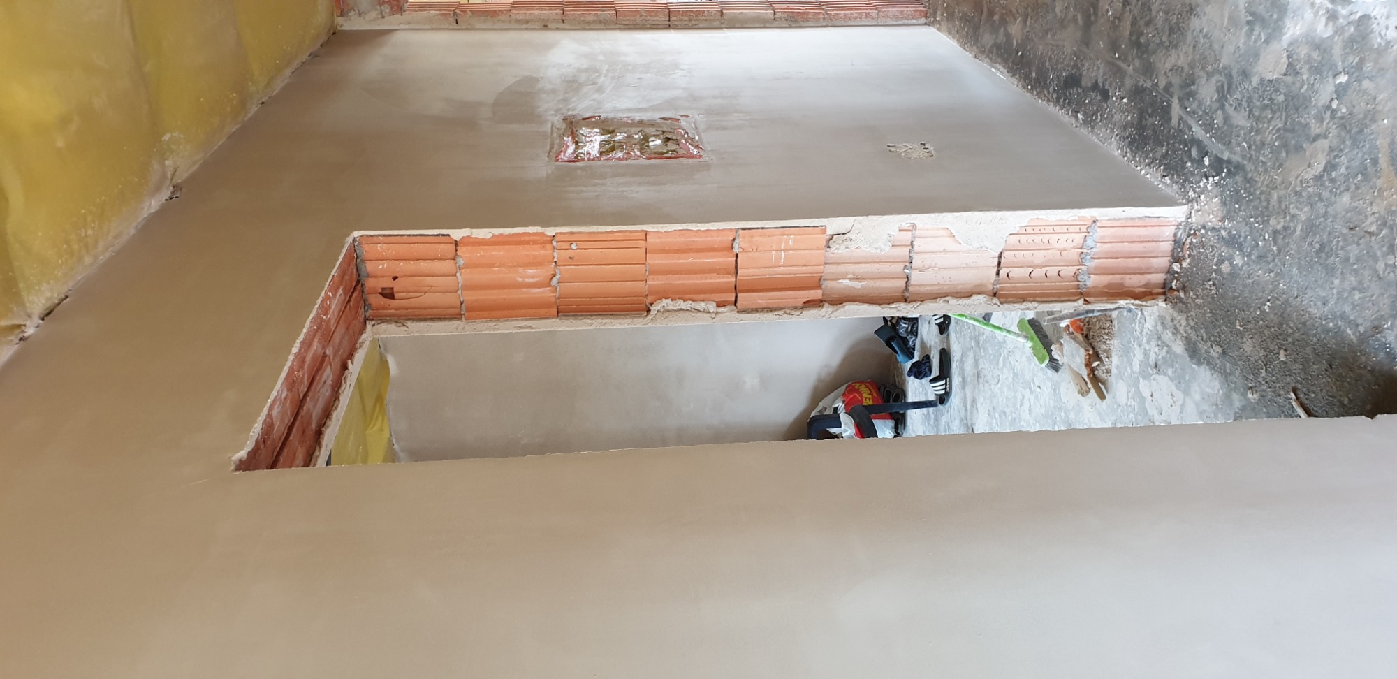 Wnętrze w trakcie prac wykończeniowych, widoczne ściany po nałożeniu gładzi, z fragmentami odsłoniętej cegły i instalacji elektrycznej, na podłodze narzędzia i torba robocza.