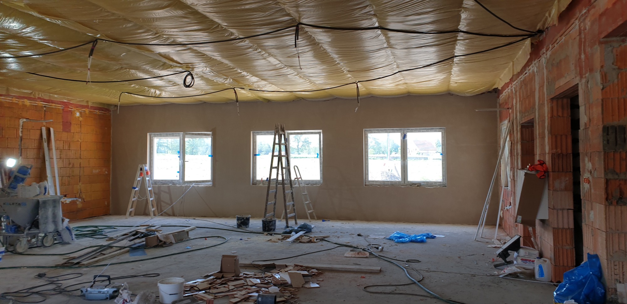 Wnętrze w trakcie szpachlowania: sufit zabezpieczony folią, ściany częściowo pokryte gładzią, widoczne drabiny, maszyna do nakładania gładzi, narzędzia i resztki materiałów na podłodze.