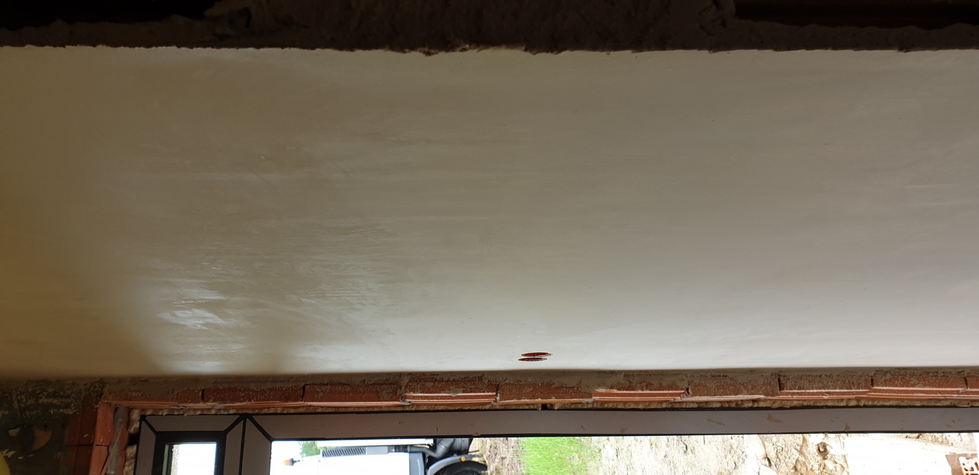 Świeżo położona, jasna gładź na suficie z widocznymi śladami szpachlowania, zarys okna w dolnej części kadru i fragmentem muru z cegieł.