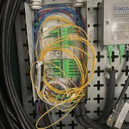 Otwarta skrzynka przyłączeniowa z gęstą plątaniną kabli światłowodowych w kolorach żółtym, niebieskim i białym, zakończonych zielonymi złączami, obok moduł optyczny Delta Electronics ONBR 1153...