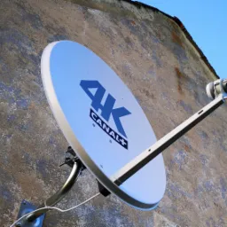 Biała antena satelitarna 4K Canal+ zamontowana na szarej, chropowatej ścianie budynku pod błękitnym niebem.