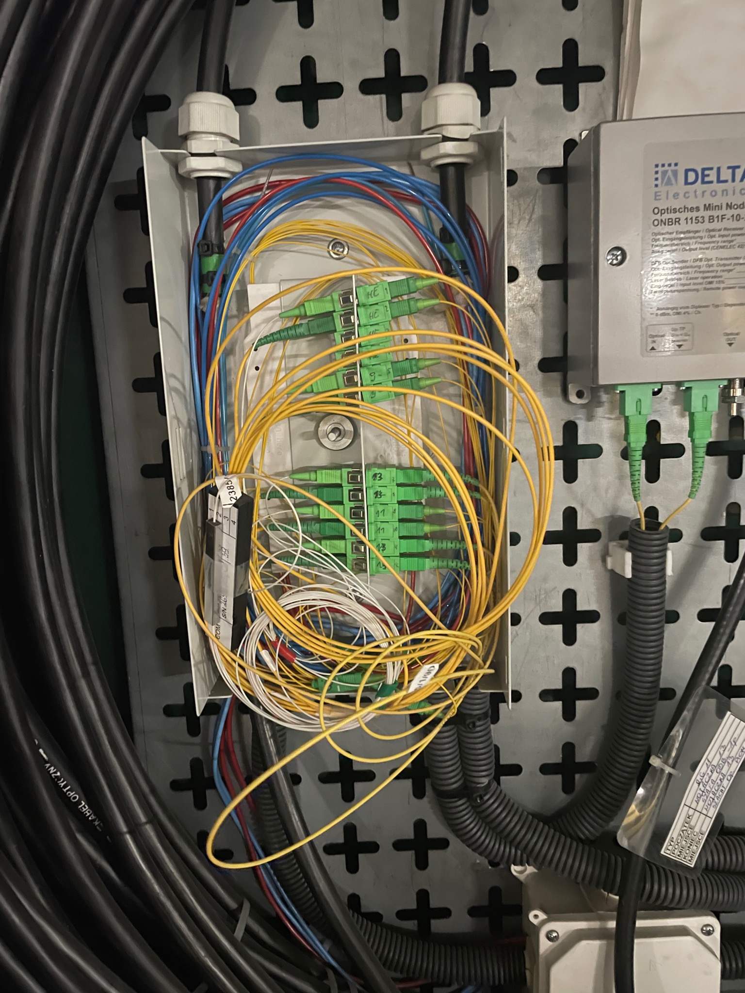 Otwarta skrzynka przyłączeniowa z gęstą plątaniną kabli światłowodowych w kolorach żółtym, niebieskim i białym, zakończonych zielonymi złączami, obok moduł optyczny Delta Electronics ONBR 1153...