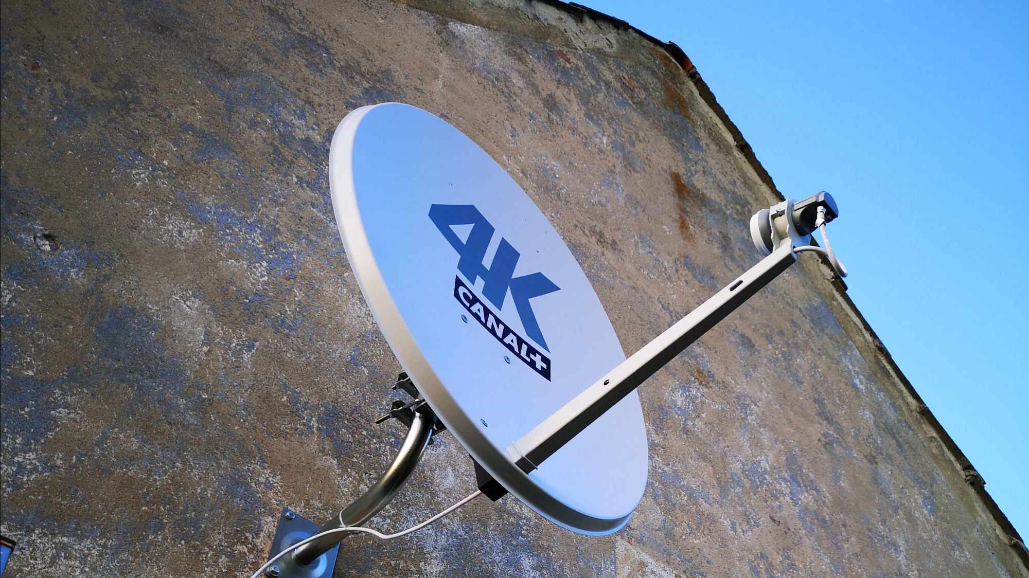 Biała antena satelitarna 4K Canal+ zamontowana na szarej, chropowatej ścianie budynku pod błękitnym niebem.