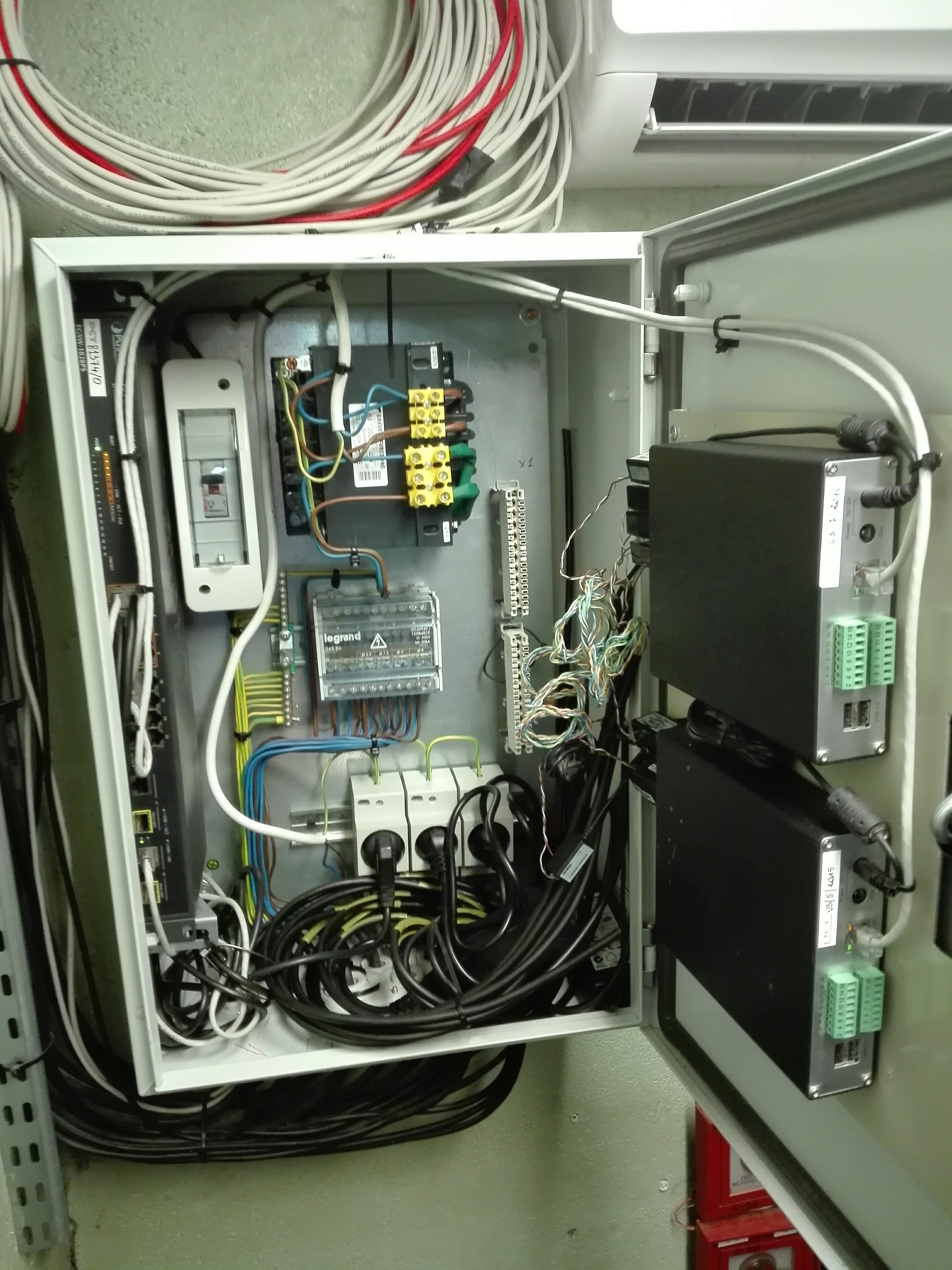 Otwarta szafa sterownicza z zamontowanymi urządzeniami elektrycznymi, kablami i złączkami; widoczne bezpieczniki, listwy zaciskowe, moduły elektroniczne z zielonymi złączami, przewody ułożone w pętle.