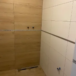 Wykończona kabina prysznicowa z drewnopodobnymi płytkami na jednej ścianie i jasnymi, gładkimi płytkami na drugiej, oddzielonymi mozaikowym paskiem. Widoczny odpływ liniowy i wystające rury...