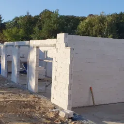 Budowa domu murowanego w trakcie wznoszenia ścian z białych bloczków, widoczne otwory okienne i drzwiowe, teren budowy z piaskiem, w tle drzewa.