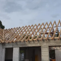 Wiązary dachowe z drewna na budynku w trakcie budowy, widoczne białe bloczki i szare nadproża, drzewa w tle