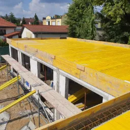 Budowa domu jednorodzinnego: widok z góry na wzniesione ściany z białego bloczka, żółte szalunki stropowe i zbrojenie wieńca, w tle zabudowa mieszkalna i zieleń drzew.