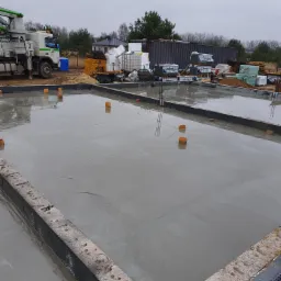 Wylany fundament z wystającymi rurami wentylacyjnymi, w tle betonowóz z pompą i materiały budowlane składowane na działce.