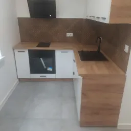 Nowoczesna kuchnia z białymi szafkami i drewnianymi blatami, wzorzysty panel dekoracyjny nad blatem, czarny okap i płyta indukcyjna, szara podłoga.