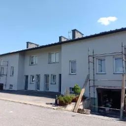 Szeregowy dom po malowaniu elewacji na szaro, z rusztowaniami i żółtym motocyklem z boku.