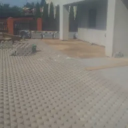 Układanie ażurowej kostki brukowej na podjeździe przed domem; widoczny pracownik w trakcie montażu, przygotowane elementy kostki oraz częściowo ułożona powierzchnia.