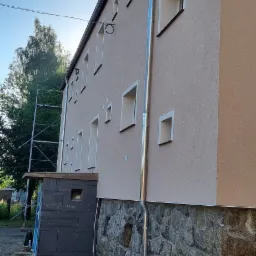 Elewacja budynku dwukondygnacyjnego z kamienną podmurówką, świeżo pomalowana na beżowo, z nową, błyszczącą rynną spustową, z widocznymi rusztowaniami w tle.