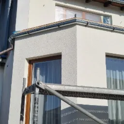 Fragment elewacji domu z widocznym rusztowaniem, okna zabezpieczone folią malarską, świeżo nałożony tynk strukturalny.