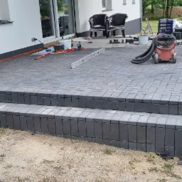 Ułożony taras z kostki brukowej z dwoma stopniami, widoczny odkurzacz przemysłowy, poziomica, paleta z kostką i stos drewna w tle.