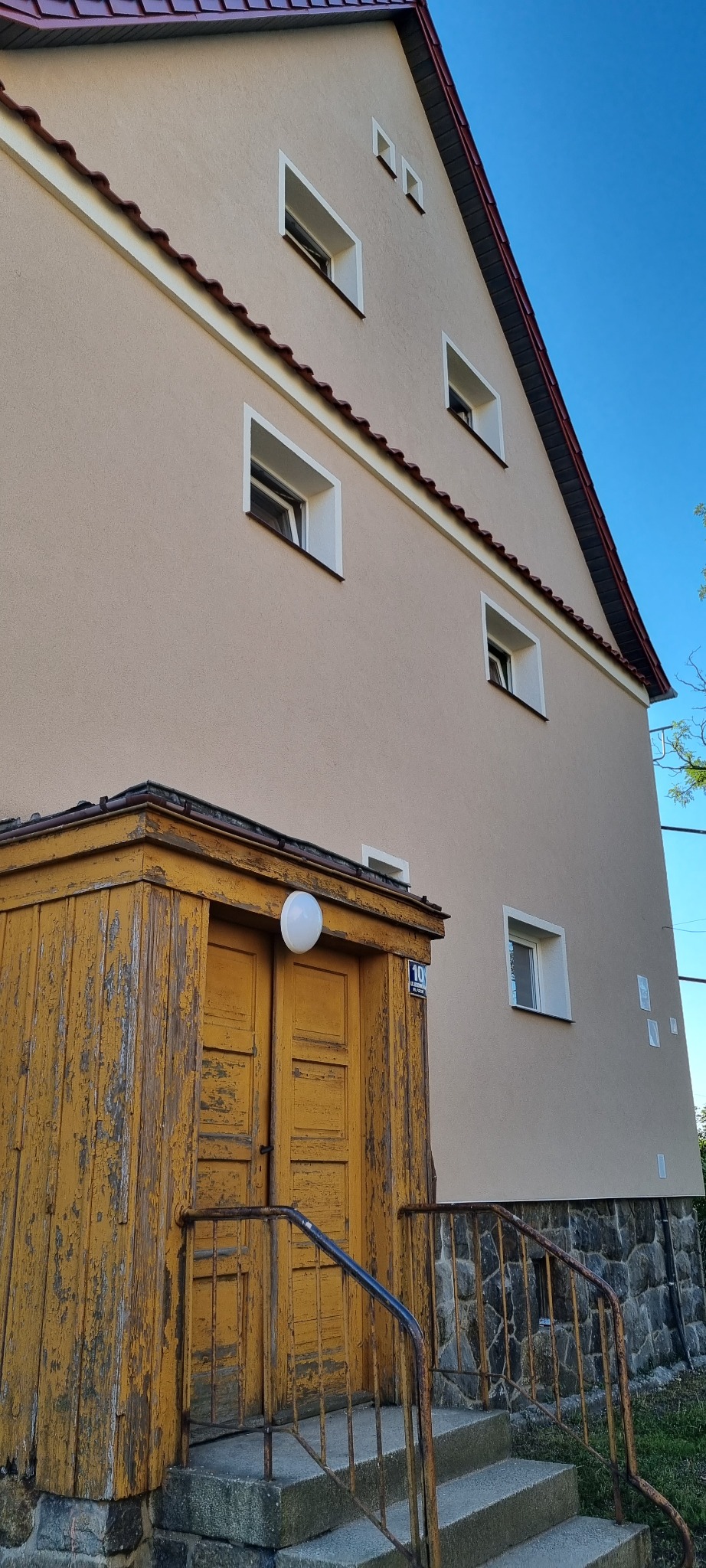 Beżowa elewacja domu z czerwonym dachem, białymi obramowaniami okien i zniszczonym, żółtym wejściem z oświetleniem kulistym oraz kamienną podmurówką i schodami.