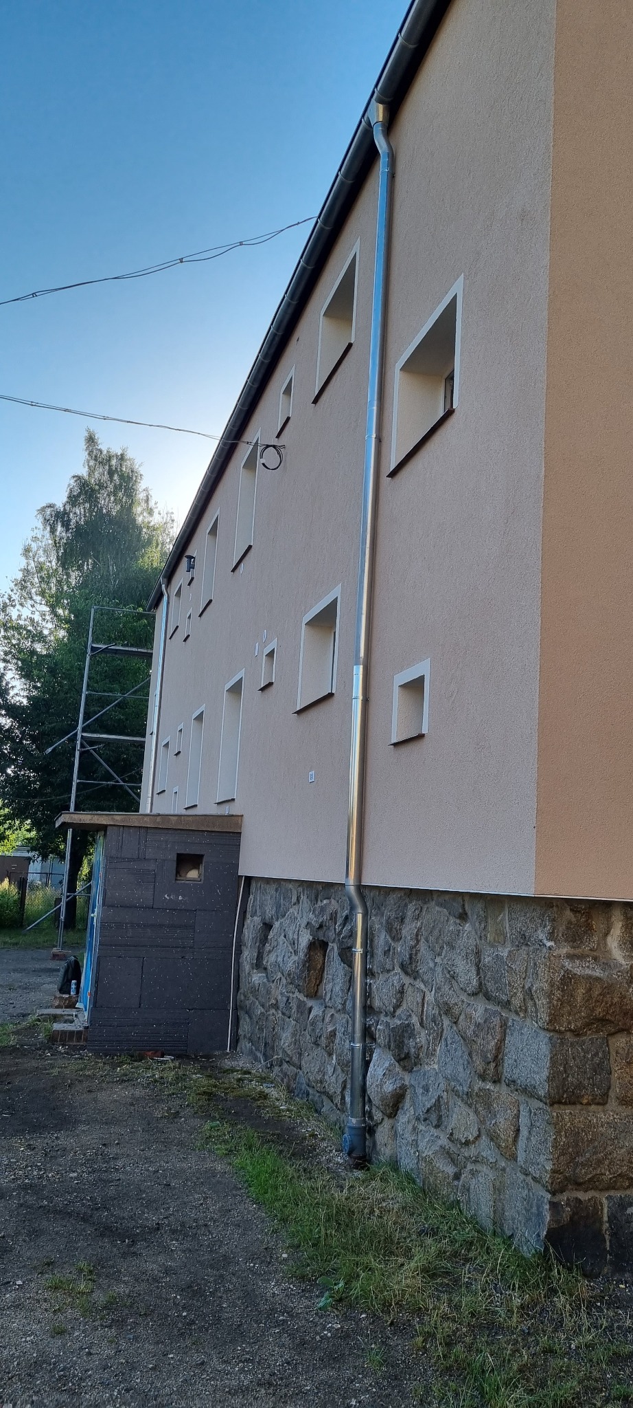 Elewacja budynku dwukondygnacyjnego z kamienną podmurówką, świeżo pomalowana na beżowo, z nową, błyszczącą rynną spustową, z widocznymi rusztowaniami w tle.