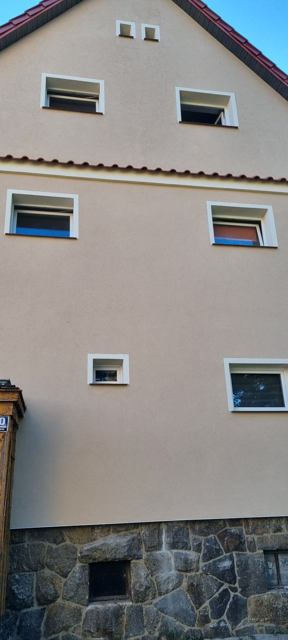 Elewacja budynku z beżowym tynkiem, białymi obramowaniami okien i kamienną podmurówką, widoczne okna o różnych rozmiarach i kształtach, część okna otwartego.