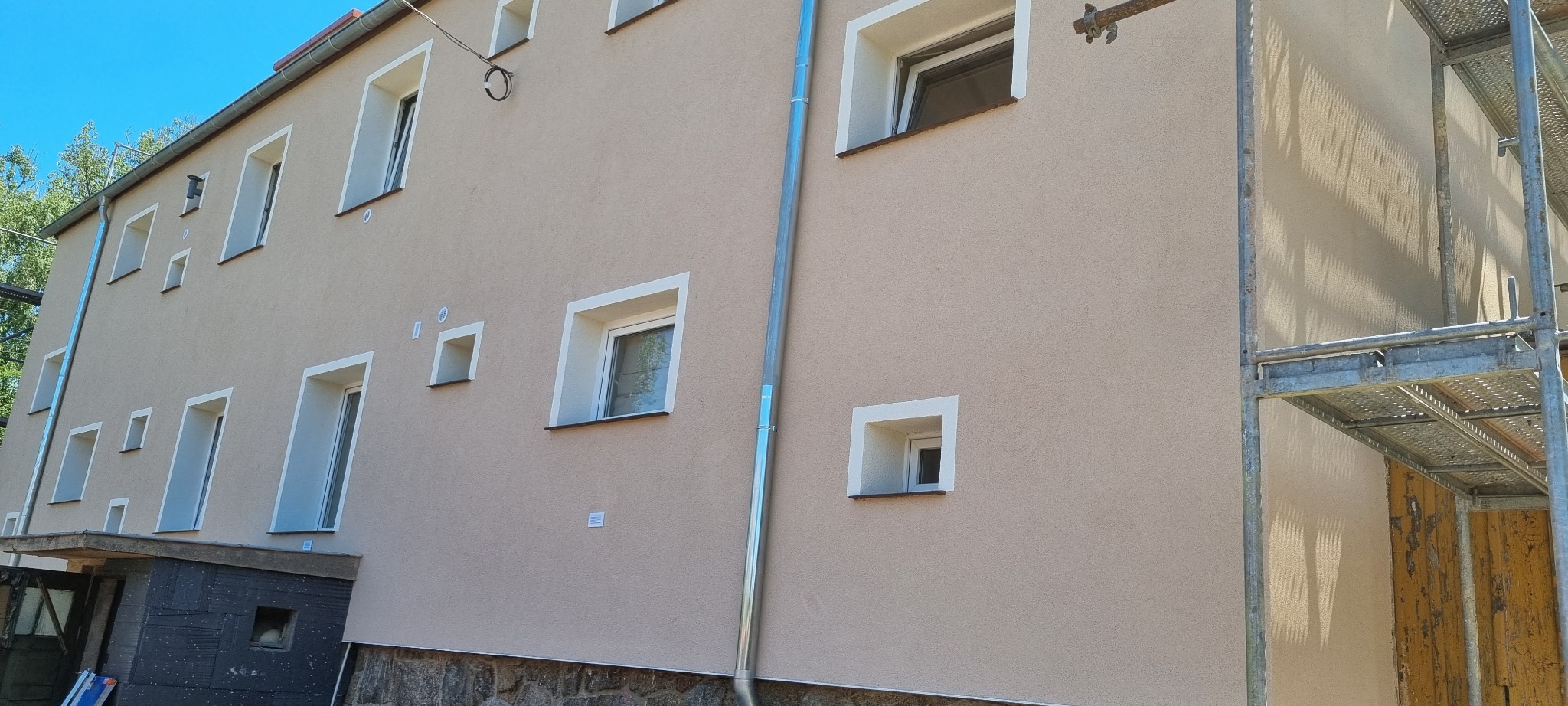 Ściana budynku po nałożeniu jasnobeżowego tynku, z białymi obramowaniami okien i stalową rynną, fragment rusztowania po prawej stronie.