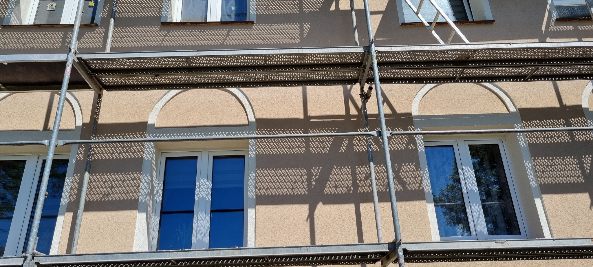 Elewacja budynku z beżowym tynkiem, białymi obramowaniami okien i łukowymi dekoracjami, częściowo zasłonięta rusztowaniem, na której widoczne są cienie