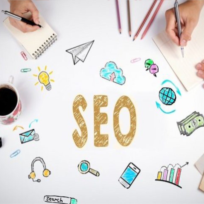 Dwie ręce piszą na notatnikach otoczonych rysunkami, w centrum napis SEO, u góry kredki kolorowe, widok z góry na białe tło.