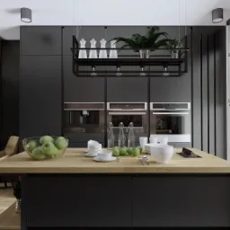 Nowoczesna kuchnia w odcieniach szarości z drewnianym blatem wyspy, na której leżą zielone jabłka, szklane butelki i biała zastawa. Nad wyspą zawieszona metalowa półka z roślinami i ceramicznymi...