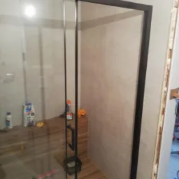 Nowoczesna kabina prysznicowa z czarną ramą i drewnianym siedziskiem wewnątrz, widoczne narzędzia i chemia budowlana, płytki w odcieniach beżu na ścianach.