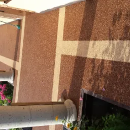 Brązowa, teksturowana powierzchnia tarasu z jasnymi pasami tworzącymi wzór krzyża, widoczne kolumny, kwiaty w doniczkach i cień drzewa.