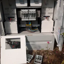 Otwarta skrzynka elektryczna z widocznymi bezpiecznikami, przewodami i narzędziem (wkrętarka akumulatorowa Bosch) wewnątrz; obok leżą elementy obudowy i rośnie róża.