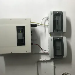 Biały inwerter firmy Sofar Solar zamontowany na ścianie obok dwóch szarych skrzynek z bezpiecznikami. Widoczne kable i rury instalacyjne.