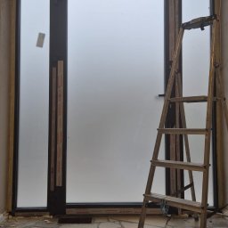 SYLWIA NAJDA - Montaż okna z matową szybą w surowym wnętrzu. Widoczna drabina, fragment podłogi z płytkami i ślady po pracach budowlanych. Okno z czarną ramą.